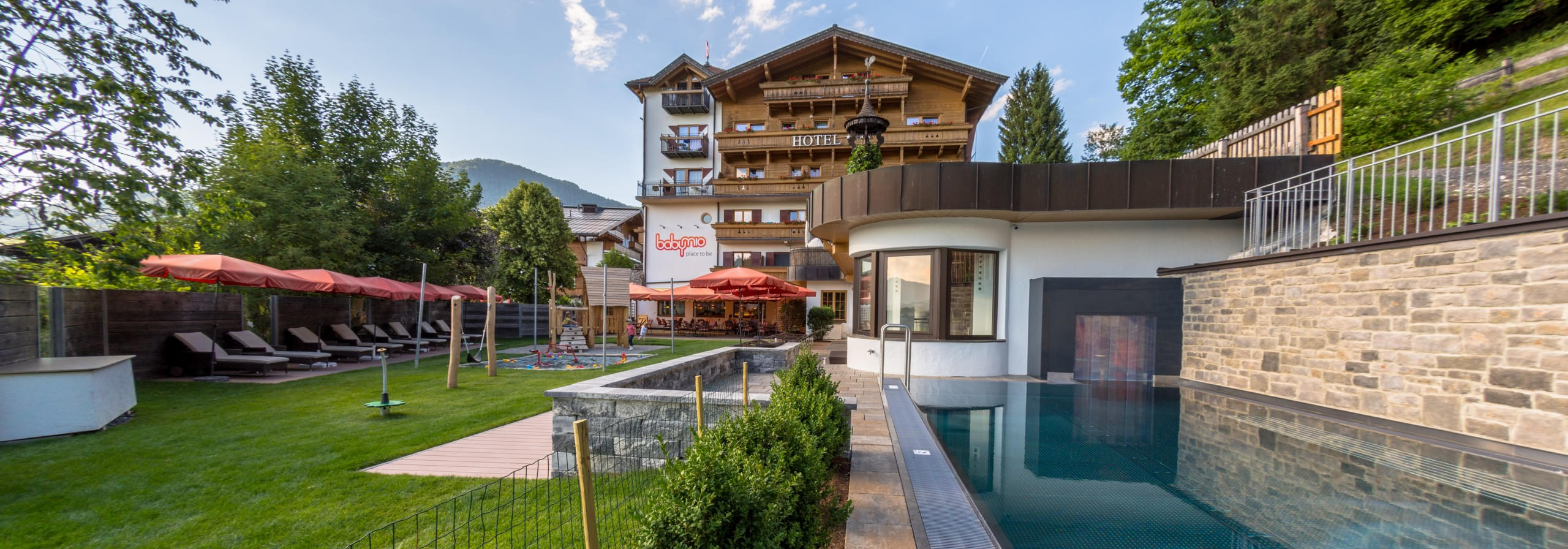 Außenansicht vom Familienhotel Babymio mit Außenpool, Gartenbereich und Spielplatz im Sommer