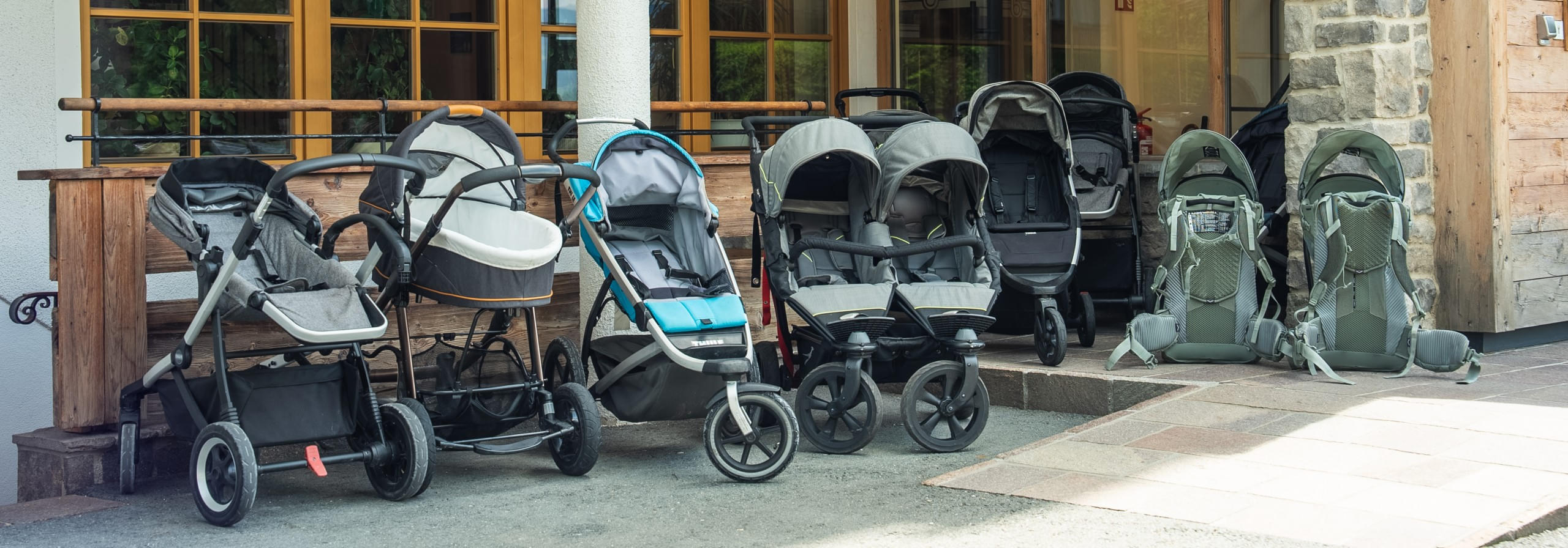 Reihe von verschiedenen Kinderwagen und auch Kindertragerucksack vor dem Hotel Babymio in Tirol
