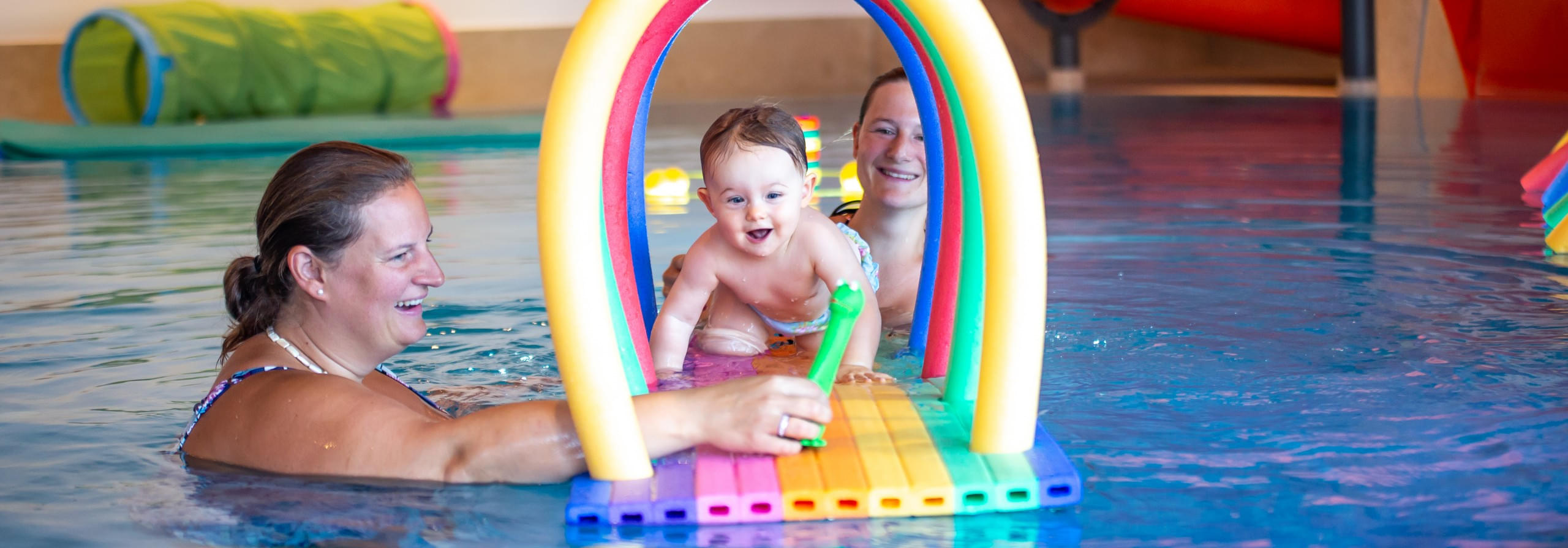 Eltern und Baby spielen im Schwimmbad mit buntem Wasserbogen