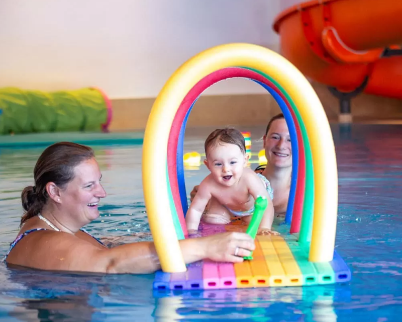 Baby schwimmt mit Hilfe von Erwachsenen unter einem bunten Schwimmbogen