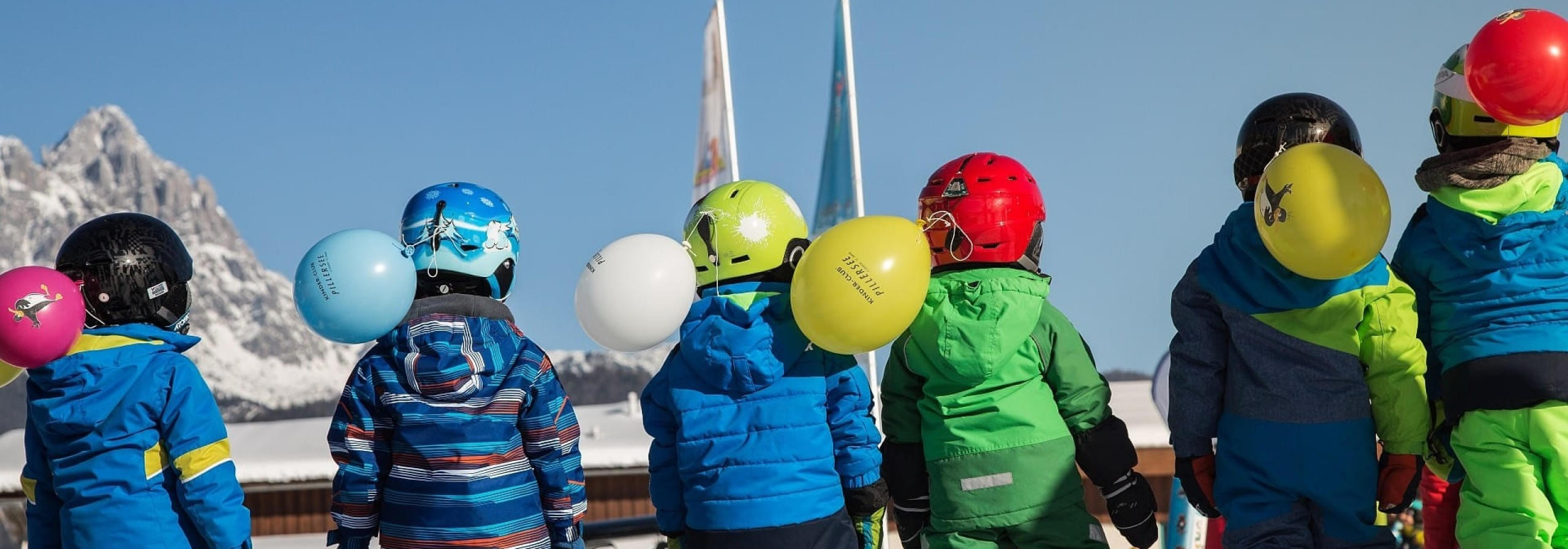 Kinder im Skianzug mit Helmen und Luftballons vor verschneiten Bergen der Kitzbüheler Alpen
